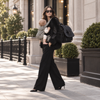 Mother walking down a city street with her baby in a houndstooth carrier, wearing the NAEVII Elara Diaper Bag Backpack in black over one shoulder#Color_Black