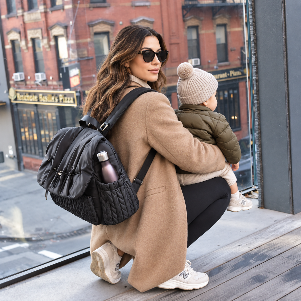  Mother in a camel coat holding her toddler while wearing the NAEVII Elara Diaper Bag Backpack in black, with a water bottle in the side pocket, by a city window#Color_Black