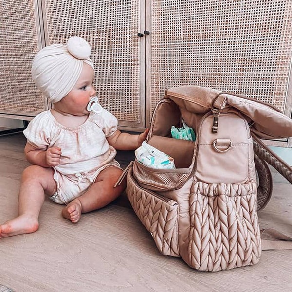 Baby in a headwrap with a pacifier sitting on the floor next to an open NAEVII Elara Diaper Bag Backpack in pink with diapers inside#Color_Pink