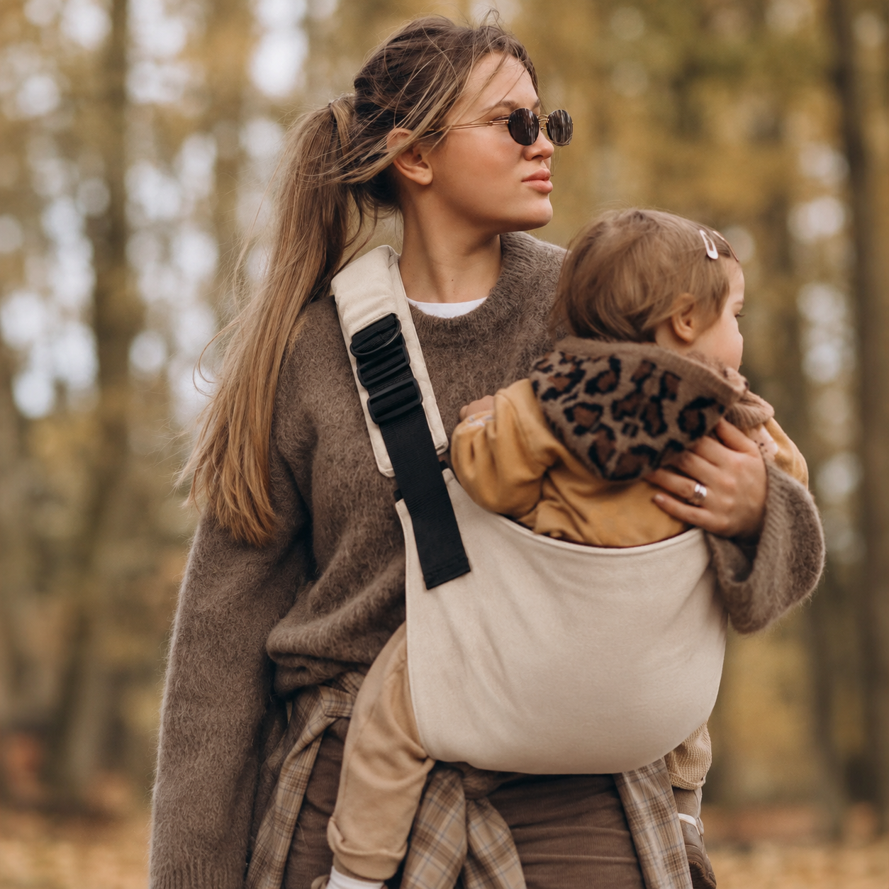 Woman carrying her toddler in the NAEVII Toddler Sling Carrier in beige, front carry on an autumn forest walk#Color_Beige
