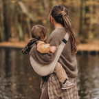 Woman seen from behind by a lake carrying her toddler in the NAEVII Toddler Sling Carrier in beige, single strap crossing the back#Color_Beige