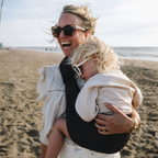Woman laughing on the beach with her toddler carried in the NAEVII Toddler Sling Carrier in black, front carry#Color_Black