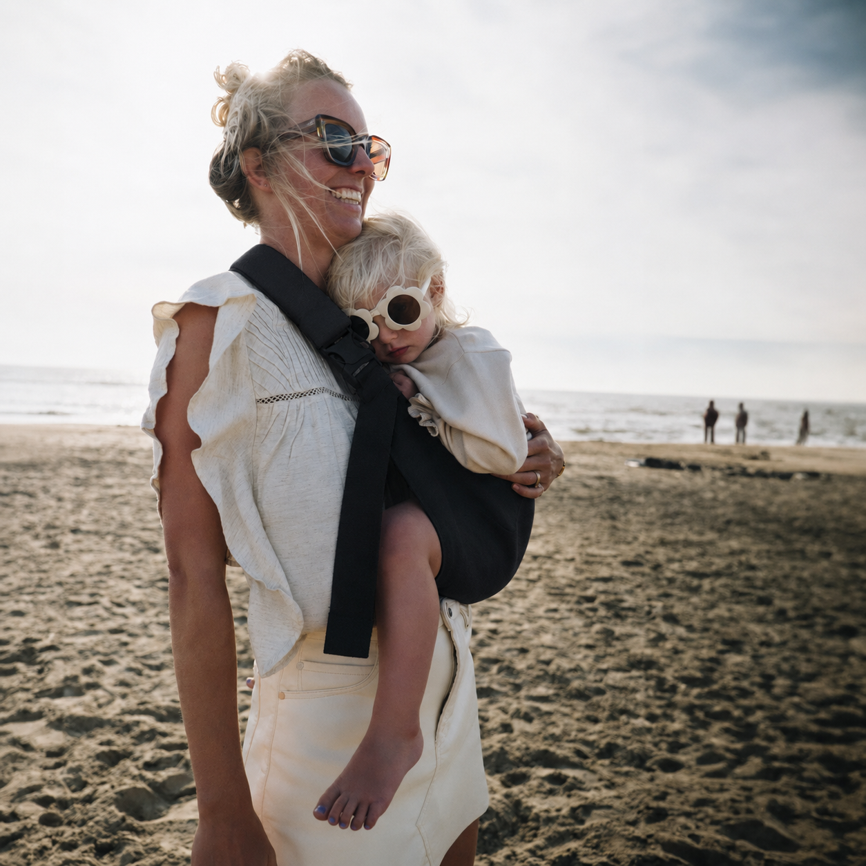 Toddler asleep against her mother in the NAEVII Toddler Sling Carrier in black, front carry on the beach#Color_Black