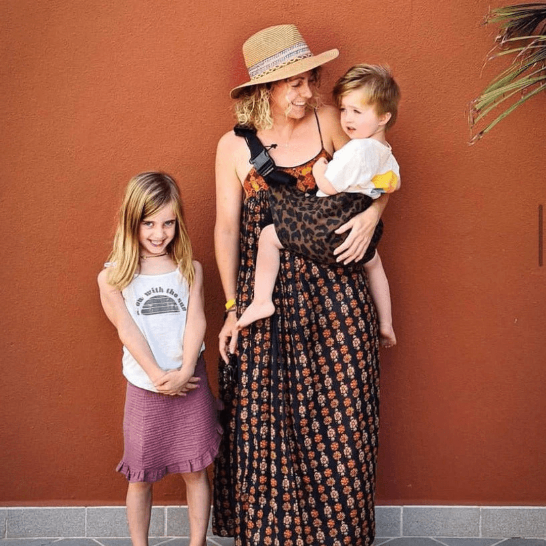 Woman carrying her toddler in the NAEVII Toddler Sling Carrier, brown leopard print, standing outdoors with her older daughter#Color_Brown Leopard