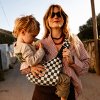 Woman carrying her toddler in the NAEVII Toddler Sling Carrier, Ebony Check, walking a sunlit village street at golden hour#Color_Ebony Check