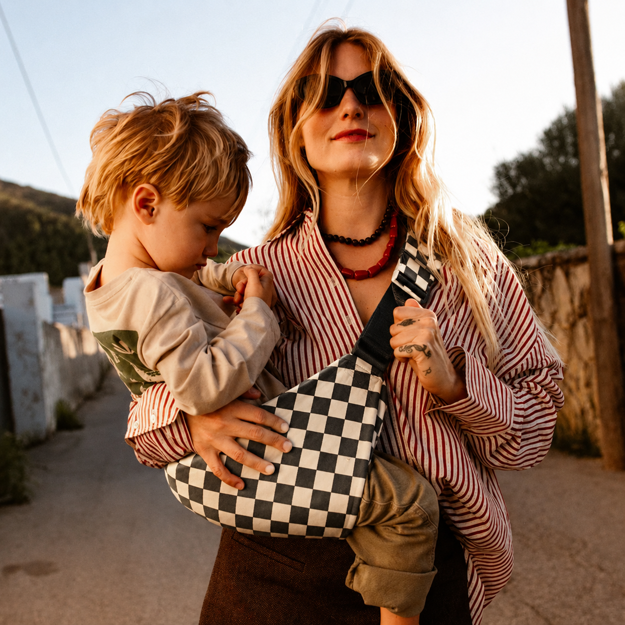 Woman carrying her toddler in the NAEVII Toddler Sling Carrier, Ebony Check, walking a sunlit village street at golden hour#Color_Ebony Check