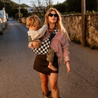 Woman walking a village street carrying her toddler in the NAEVII Toddler Sling Carrier, Ebony Check, hands free at golden hour#Color_Ebony Check