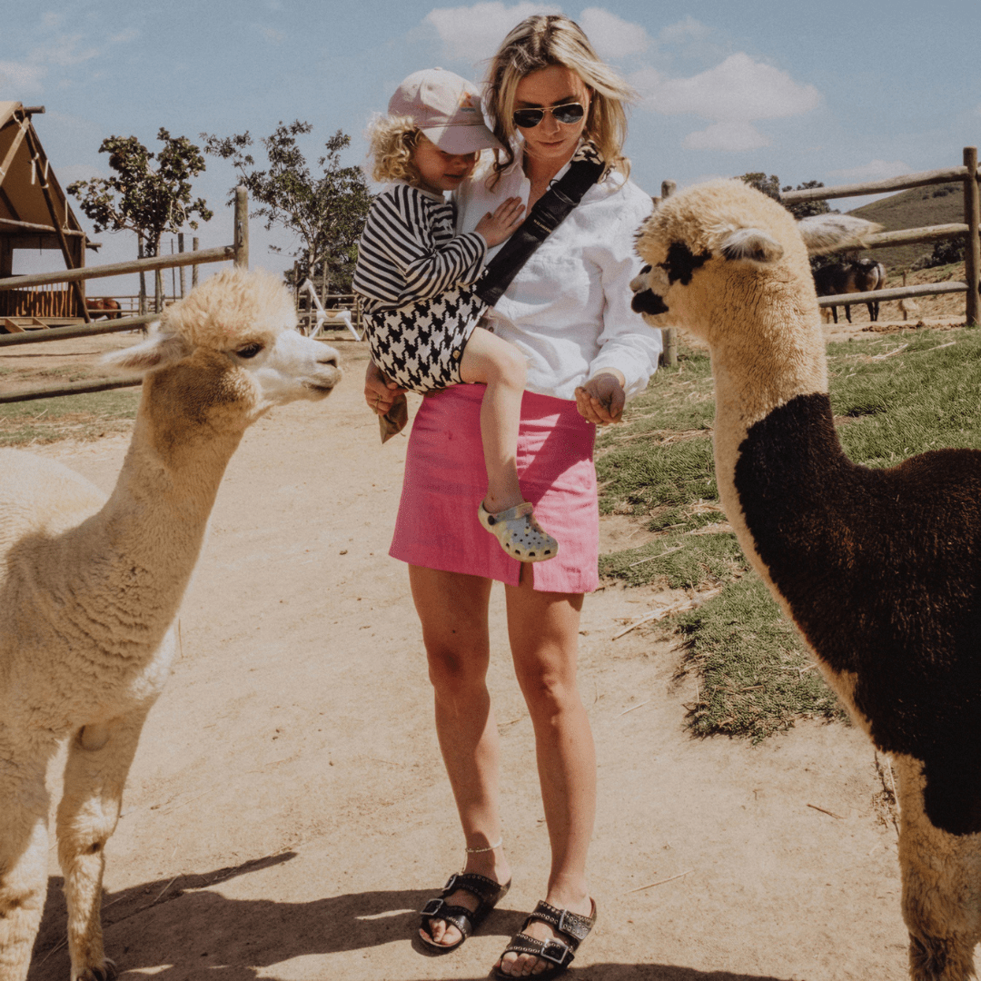 Woman feeding alpacas while carrying her toddler in the NAEVII Toddler Sling Carrier, black and white Houndstooth, outdoors at a farm#Color_Houndstooth