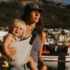Woman carrying her toddler in the NAEVII Toddler Sling Carrier, Neutral Houndstooth, front carry outdoors at a harbour#Color_Neutral Houndstooth